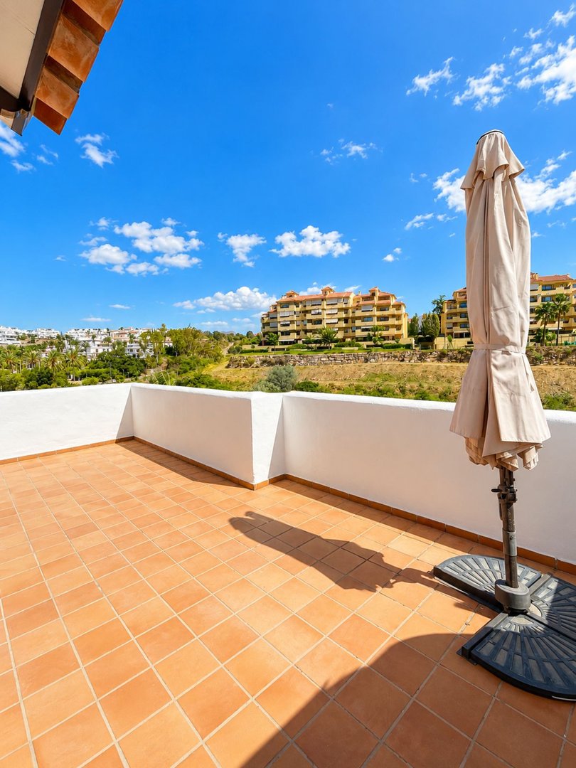 Spacious terrace with terracotta tiles and an umbrella, offering a view of resort buildings and a bright blue sky.