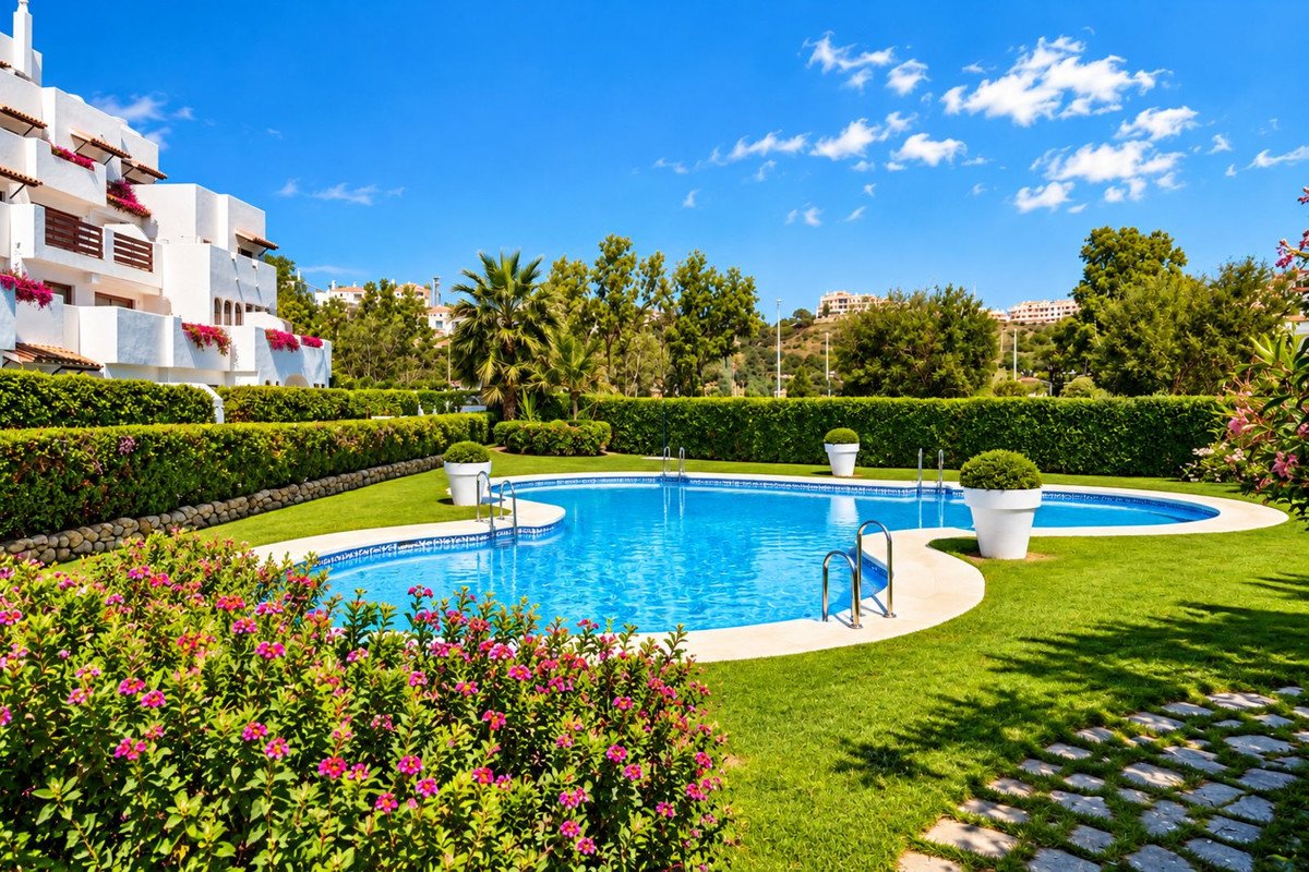 A resort swimming pool surrounded by lush green grass, manicured hedges, and flowering plants, with a white building in the background.