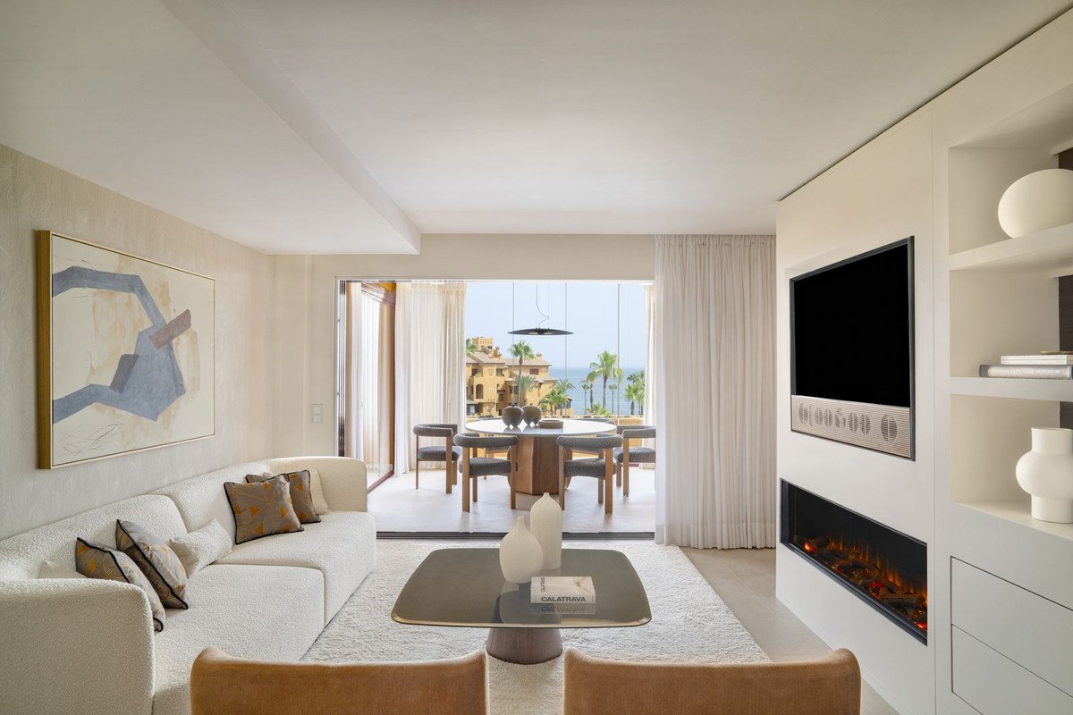 Modern living and dining area with a large white sofa, fireplace, and a balcony overlooking the sea and palm trees.