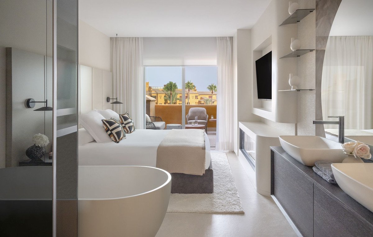 Luxury bedroom with a freestanding bathtub, king-size bed, and private balcony overlooking a resort.