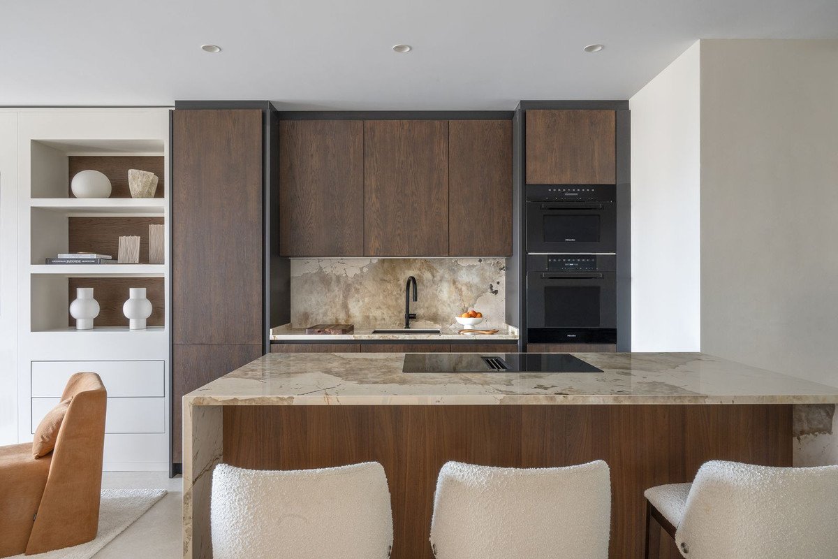 A modern kitchen with a marble island, dark wood cabinetry, built-in ovens, and a decorative backsplash.