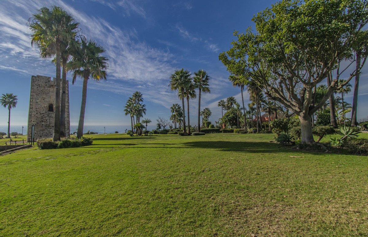 A stone tower and palm trees in a green lawn with the ocean visible in the distance under a blue sky.