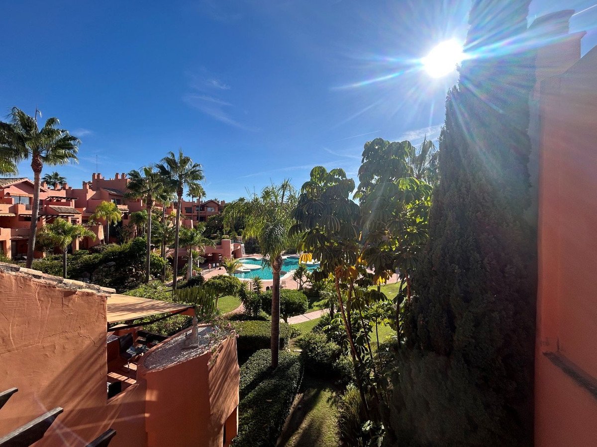 Balcony view of a resort with palm trees, a swimming pool, and Spanish-style buildings under a bright sunny sky.