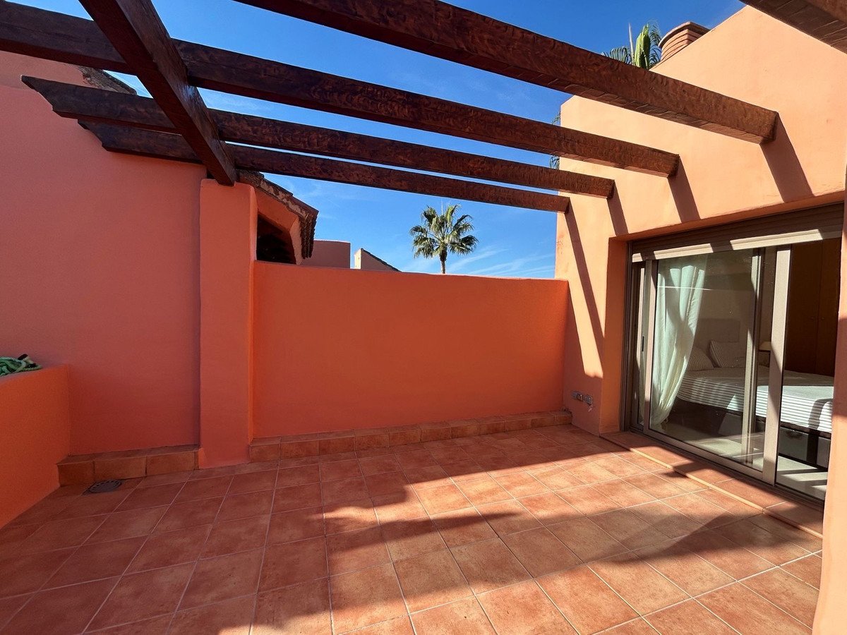 Terrace with wooden pergola and sliding glass door leading into a bedroom, with a palm tree visible against a blue sky.