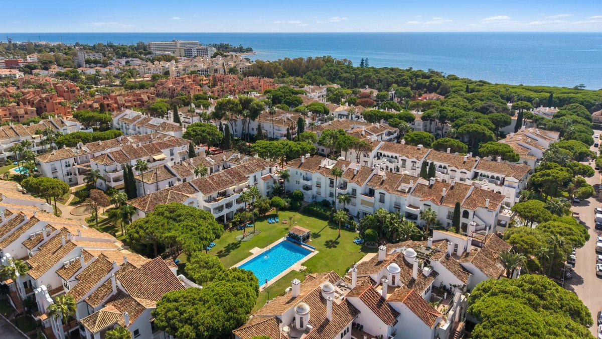 Aerial view of apartment complex with pool and green lawns, surrounded by trees and overlooking the sea on a sunny day.