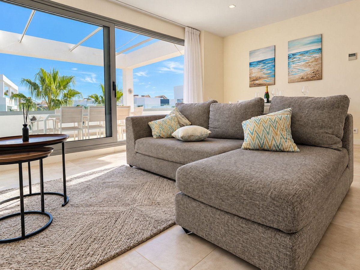 A bright living room with a grey sofa, coastal artwork, and large windows showing a sunny outdoor patio and palm trees.