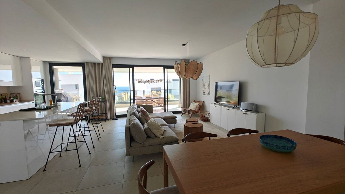 Spacious open-plan living, dining, and kitchen area with a large sectional sofa and a balcony overlooking the sea.