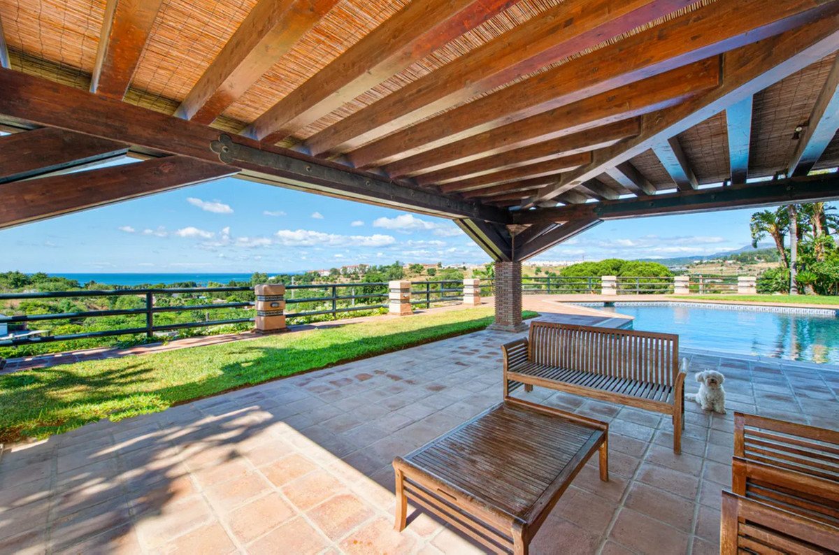 Covered wooden patio with seating and coffee table overlooking a swimming pool and the sea on a sunny day.