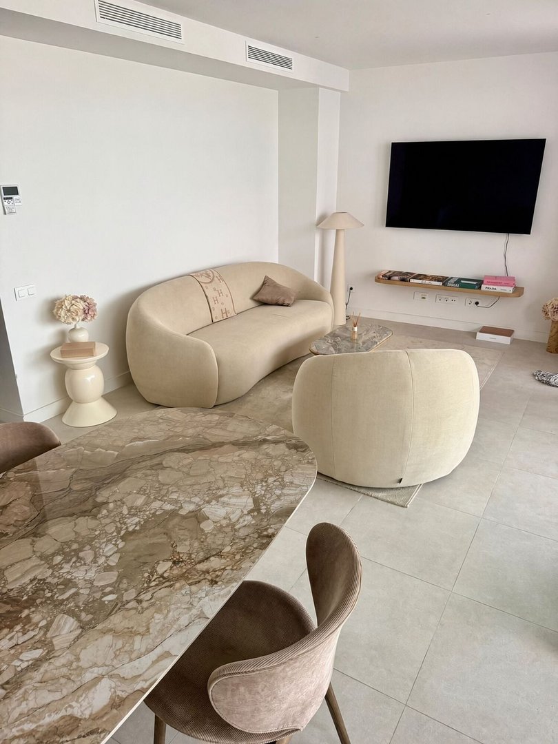 Modern living and dining area with a curved sofa, marble table, chairs, and a mounted TV.