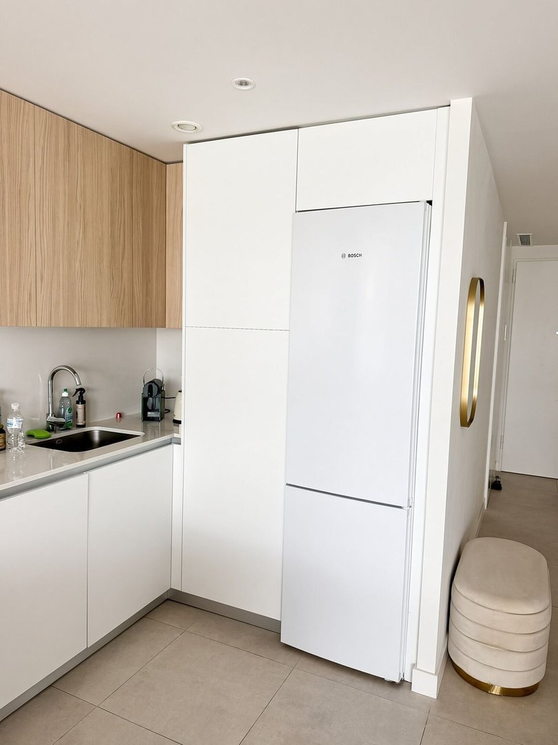 A modern kitchen with white cabinets, wooden upper cabinets, a white Bosch refrigerator, and a sink area. A beige ottoman in the corner.