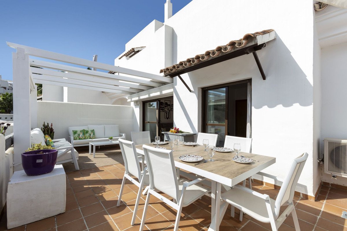 Outdoor terrace with dining table, white chairs, and a white sofa under a pergola, set for a meal.