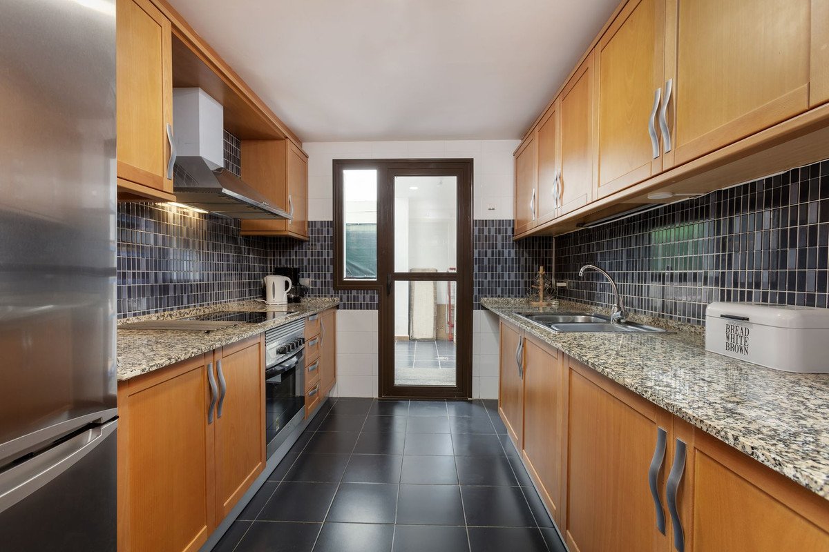 Modern galley kitchen with light wood cabinets, granite countertops, dark tile backsplash, and a stainless steel refrigerator. A glass door leads outside.