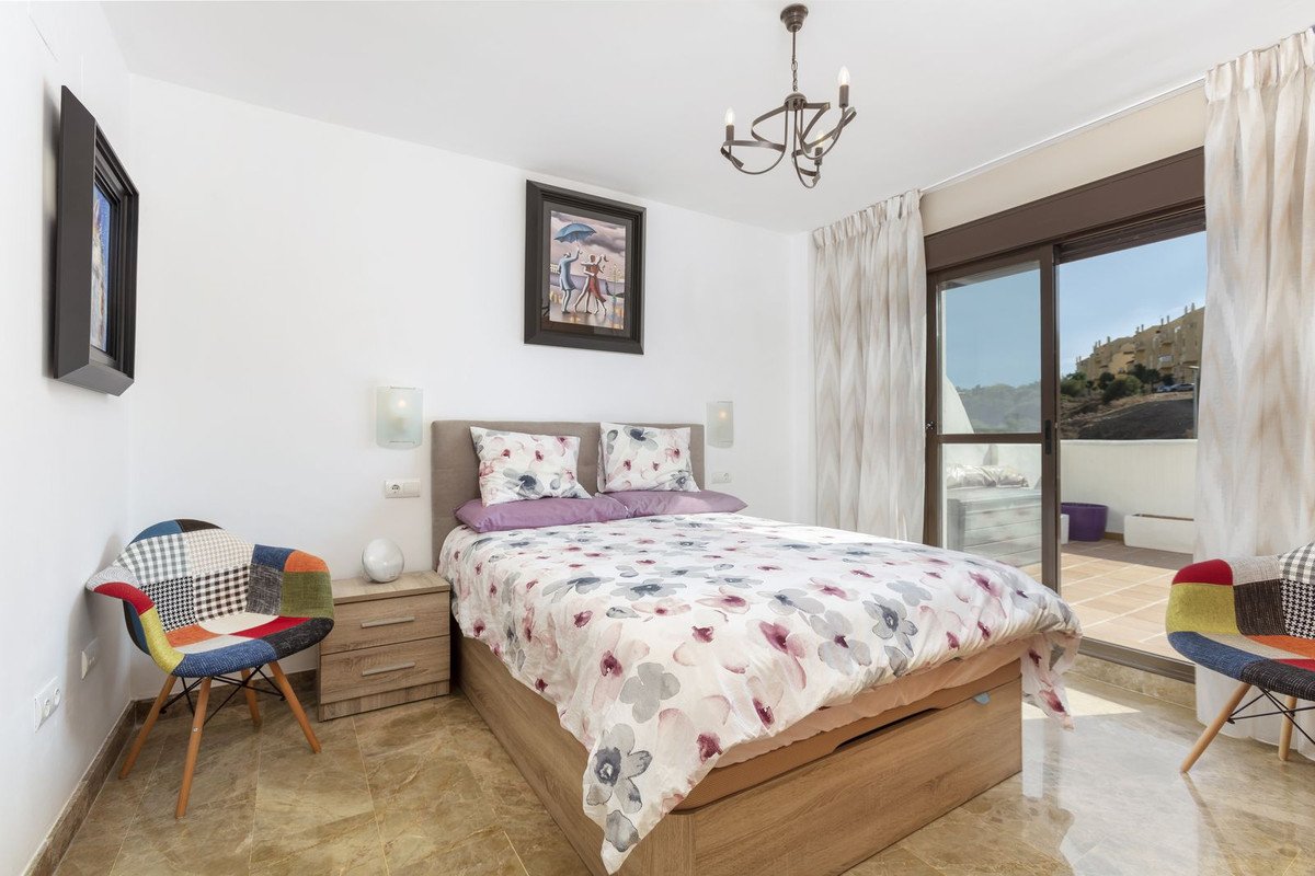 A bedroom with a double bed, floral bedding, colorful chairs, and a sliding glass door leading to a balcony with a view of hills.