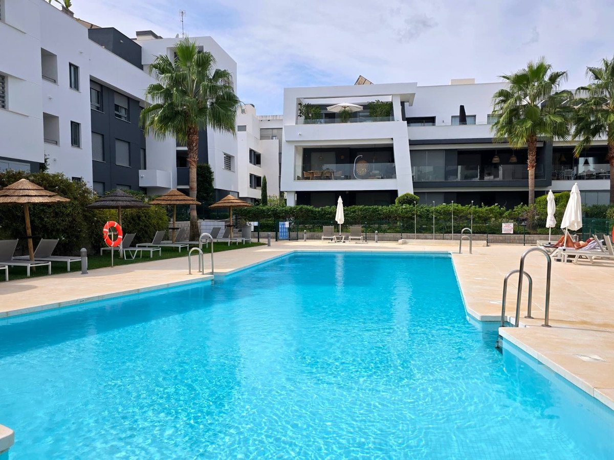 Modern apartment complex with a large blue swimming pool and sun loungers surrounded by palm trees and umbrellas.
