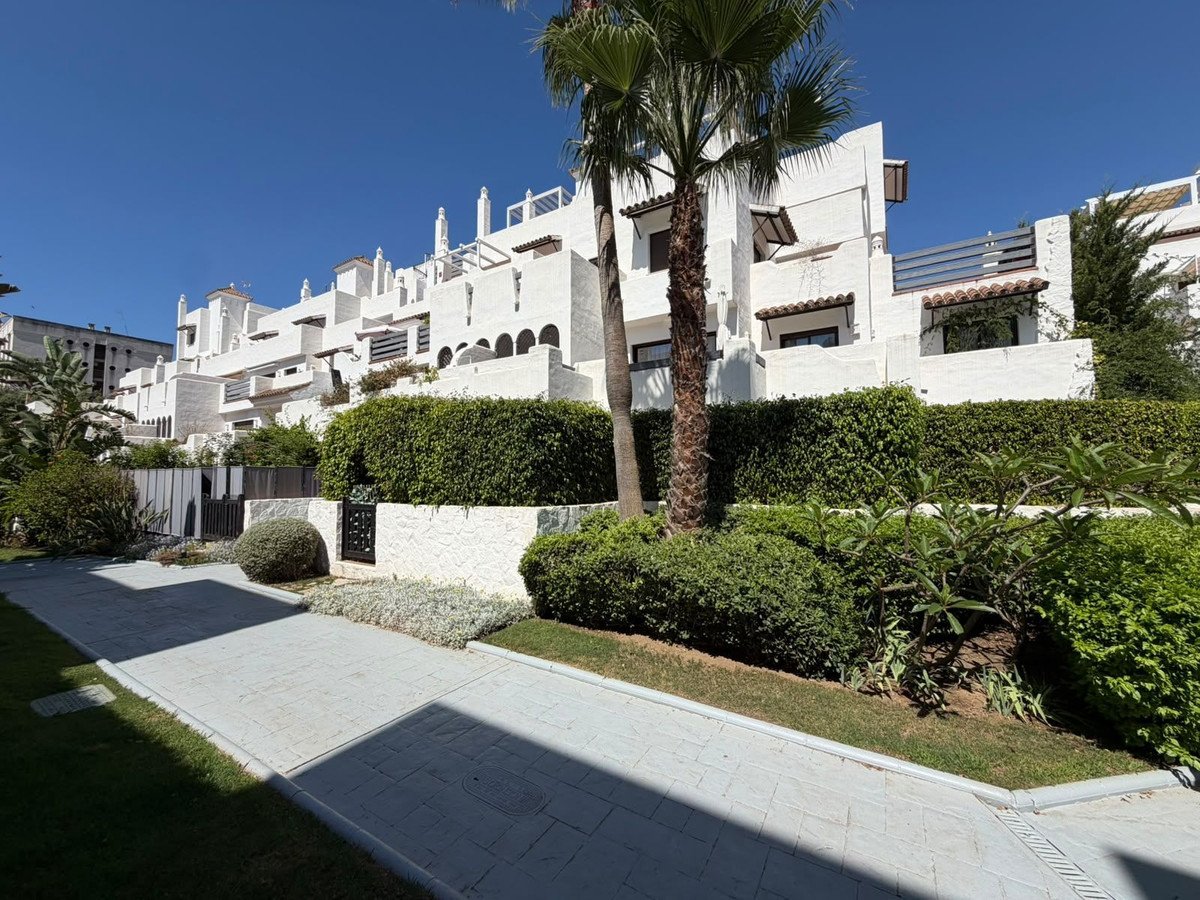 A view of a white building with multiple levels, surrounded by lush green hedges and palm trees, under a clear blue sky.