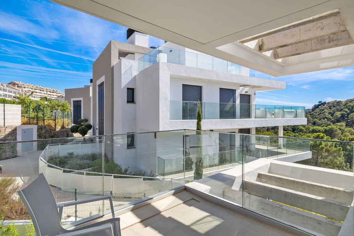 Modern villa with glass balconies and sea views, surrounded by lush green trees under a blue sky.