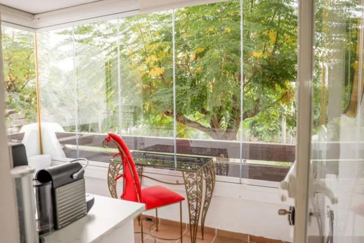 A sunroom with glass walls overlooking a large tree, featuring a red chair and a small desk.