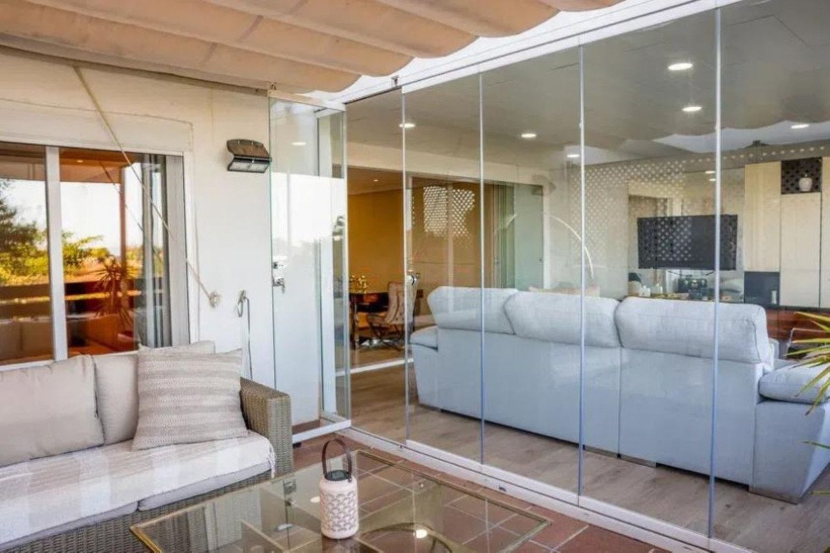 A bright sunroom with a large sofa, glass walls offering a view of the outdoors and an interior living space with a TV.