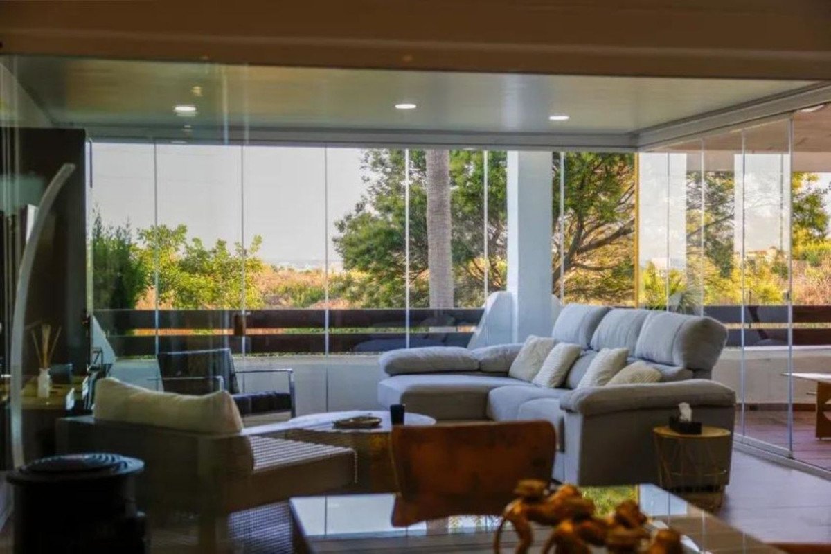 Modern living room with a large sectional sofa, floor-to-ceiling glass walls, and a scenic view of trees and landscape.