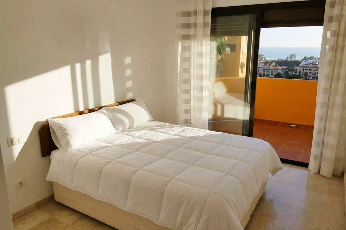 A cozy bedroom with a large white bed and a balcony offering a beautiful sea view and cityscape.