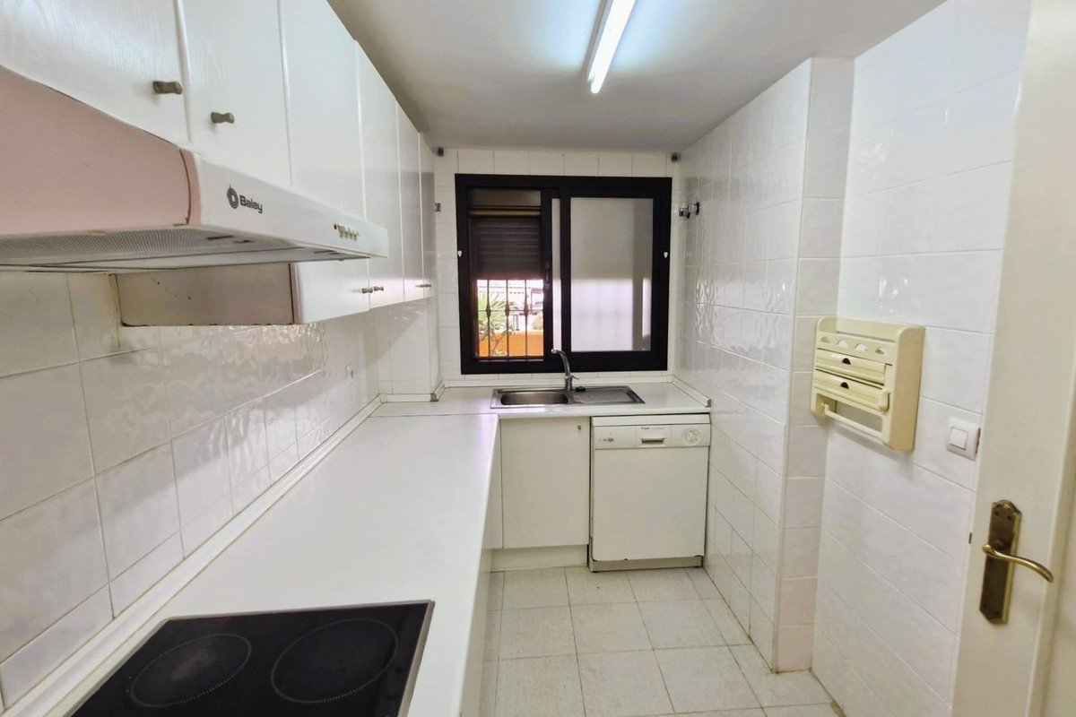 A modern white kitchen with clean cabinets, a sink, a dishwasher, and a stovetop. A window shows an exterior view.