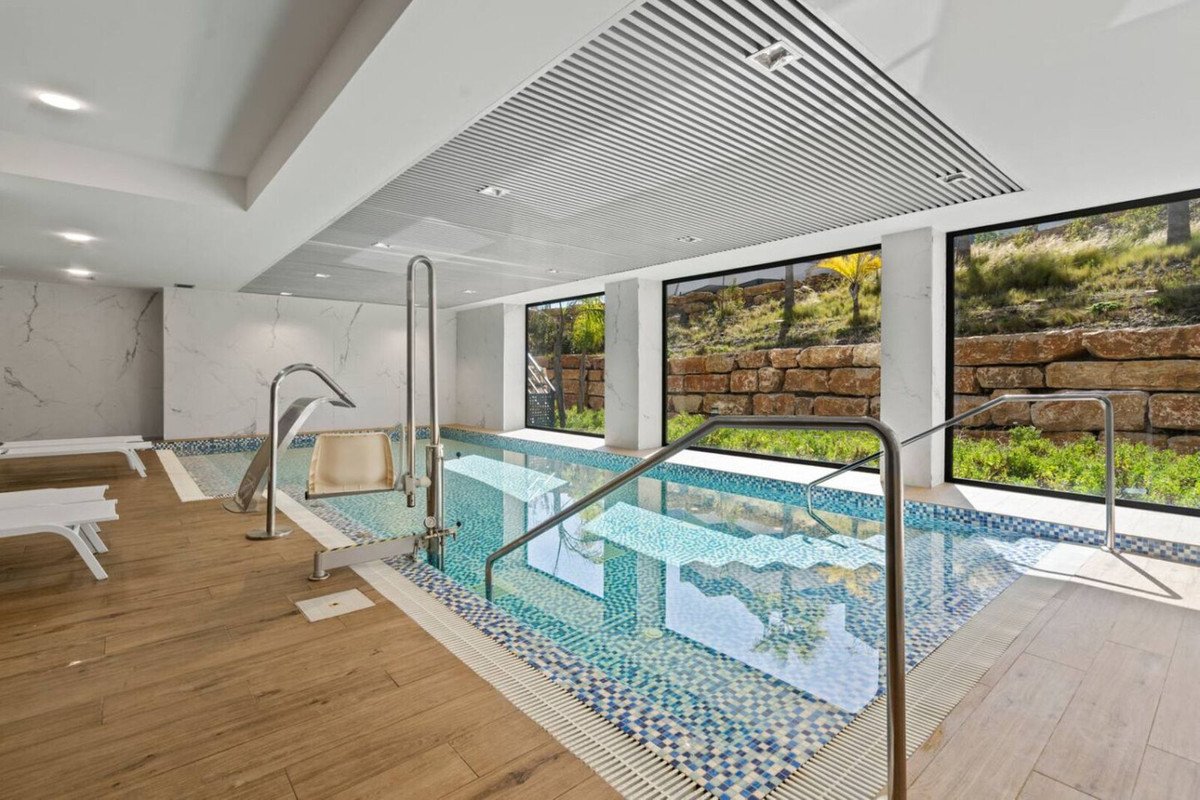 An indoor swimming pool with a water feature, steps, and large windows offering views of a stone wall and greenery.