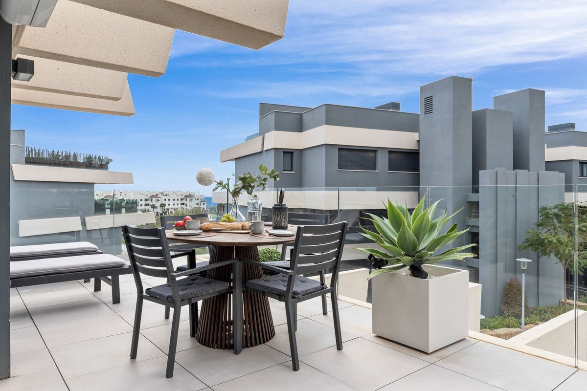 Modern terrace with dining table and chairs, offering a view of contemporary buildings and the sea in the distance.