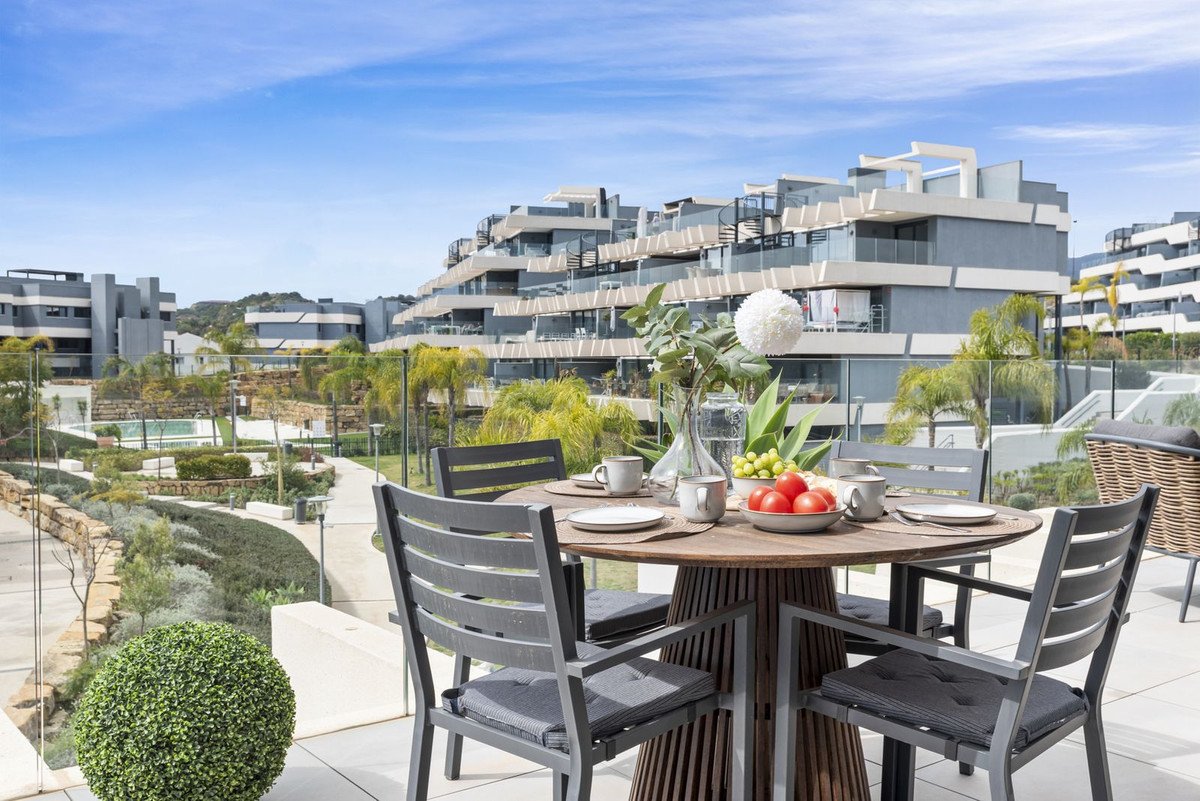 A round wooden table set for dining on a terrace, overlooking modern apartments and landscaped gardens undergrowth.