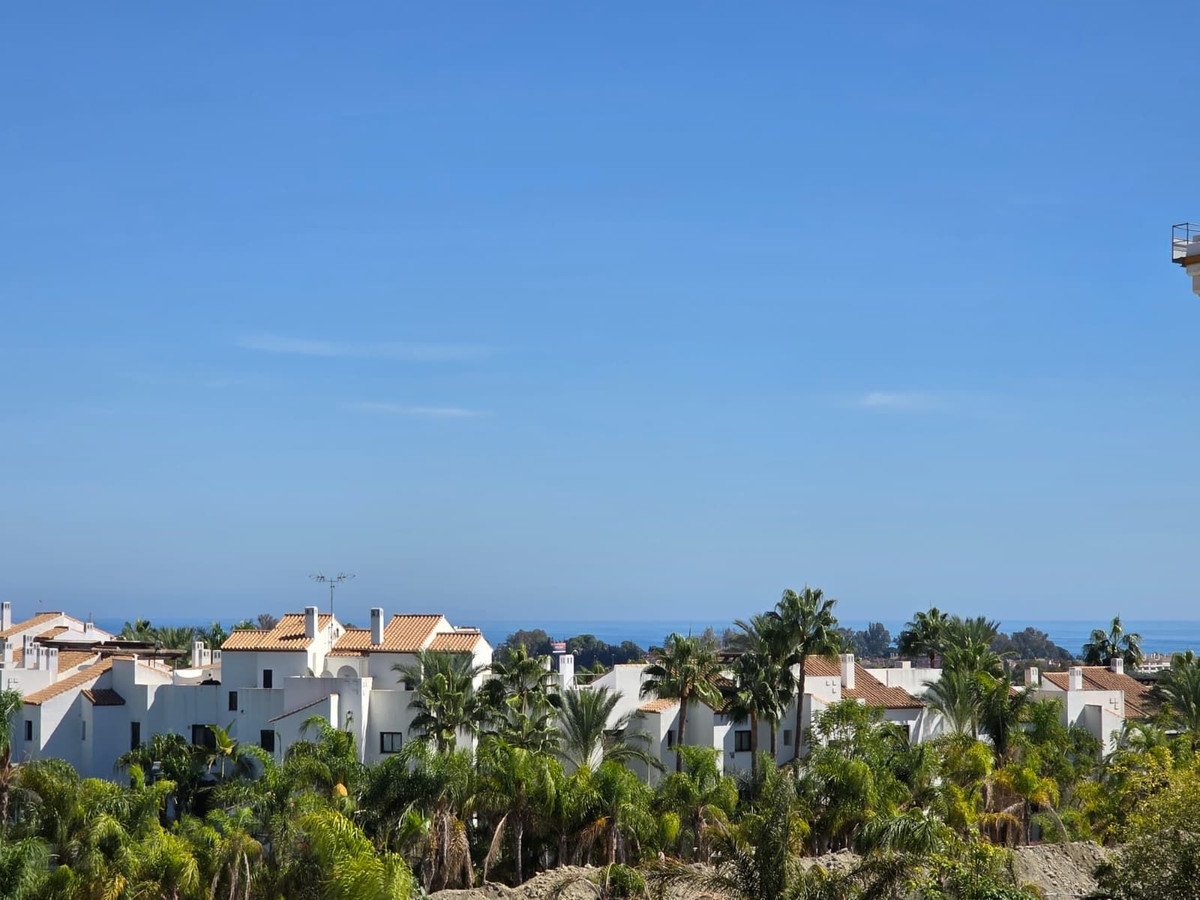 View of white Mediterranean-style houses with terracotta roofs, surrounded by palm trees, with the sea and sky in the background.