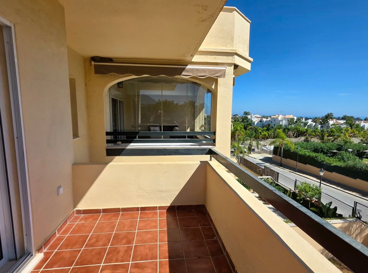 A sunny balcony with tiled floor and railing, overlooking a neighborhood with palm trees and distant sea.