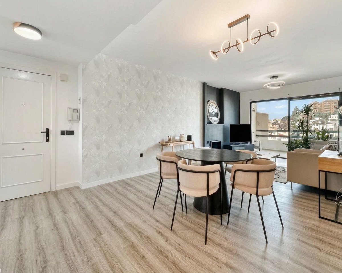 Modern dining area with a round table and chairs, leading to a living room with a view of the city and greenery.