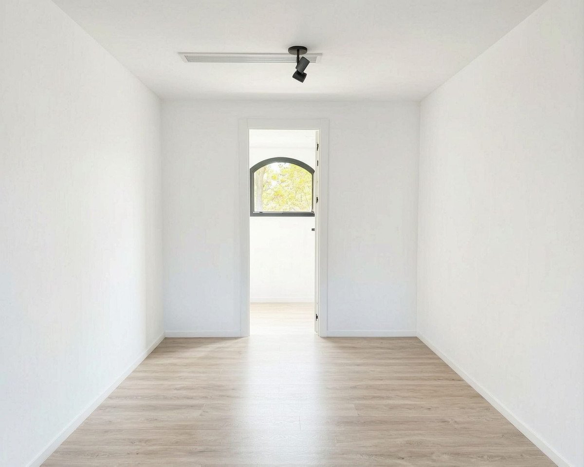 Bright empty room with white walls, wooden floor, track lighting, and an arched window with a view of trees.