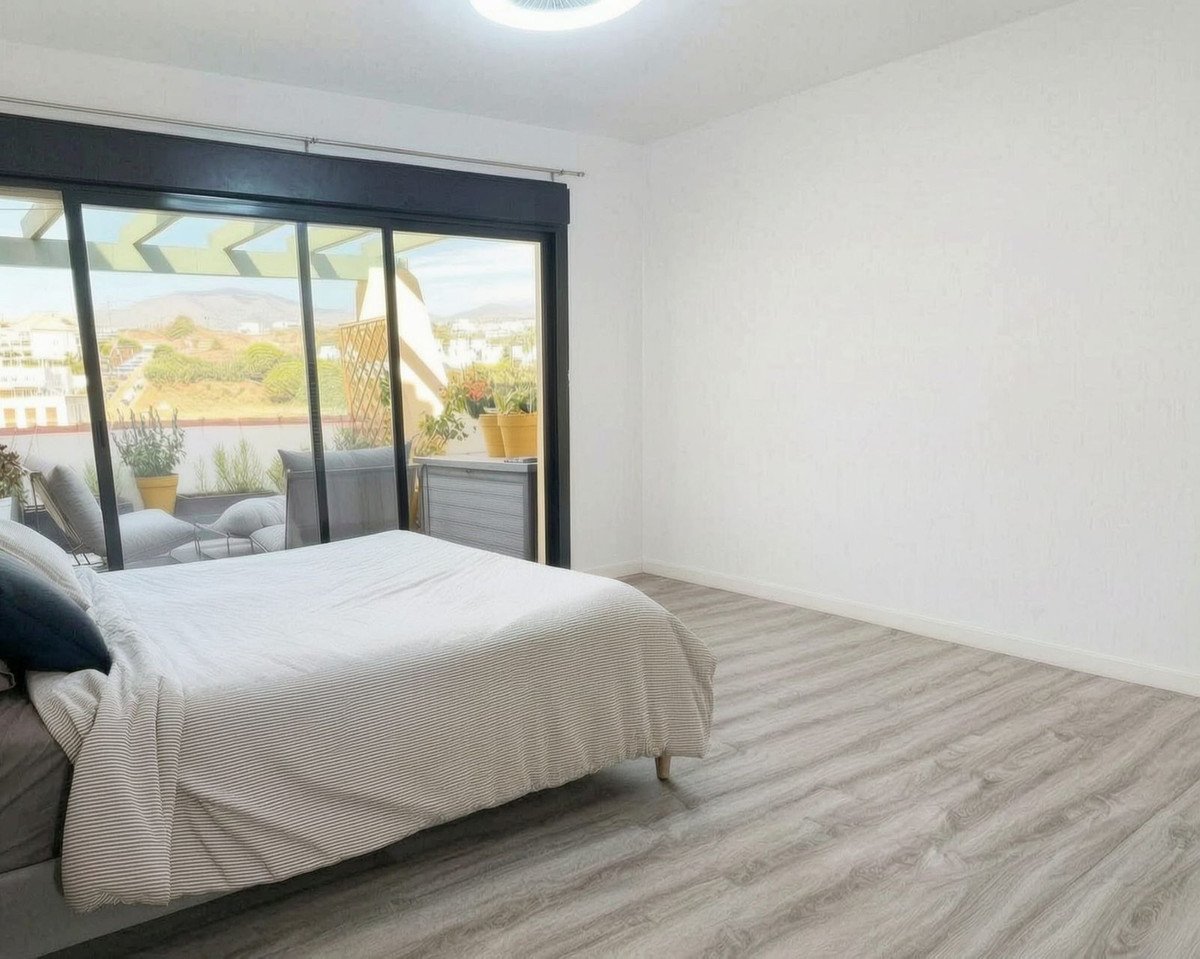 A bedroom with a large bed and sliding glass doors leading to a balcony with outdoor seating and a distant hills.