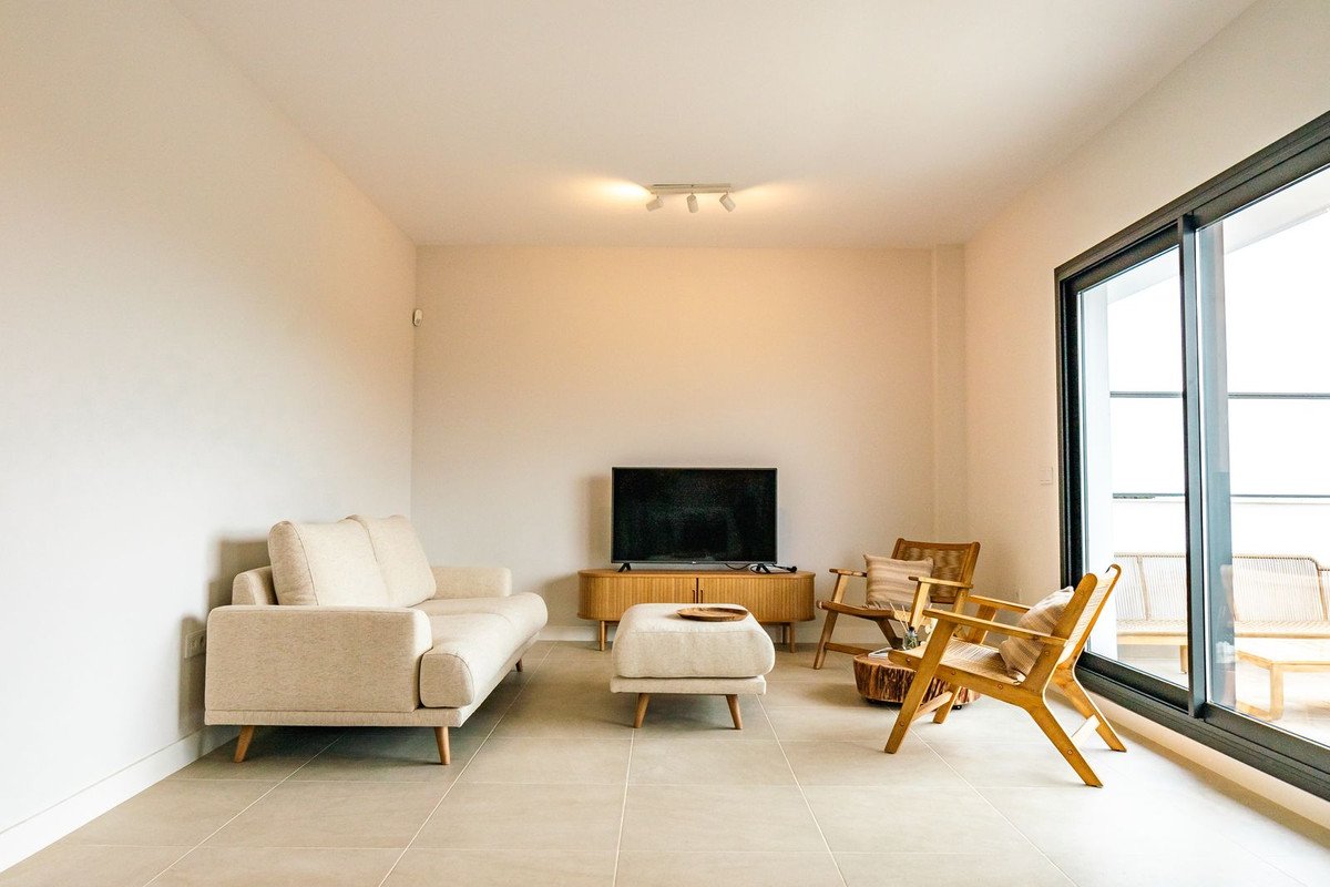 A modern living room with a sofa, chairs, ottoman, and a TV on a wooden cabinet, with sliding doors leading to an outdoor balcony.