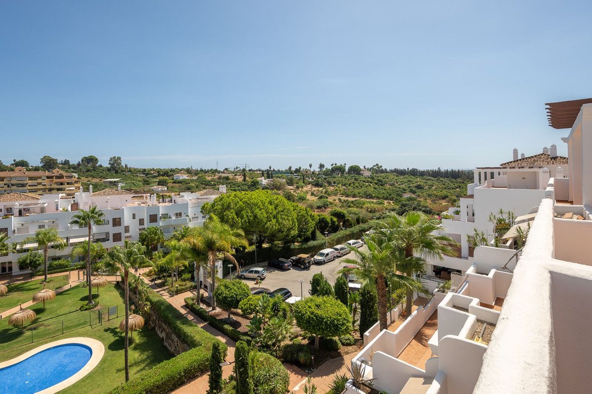 View from balcony of a complex with apartments, pool, gardens, palm trees, and distant green hills under a blue sky.