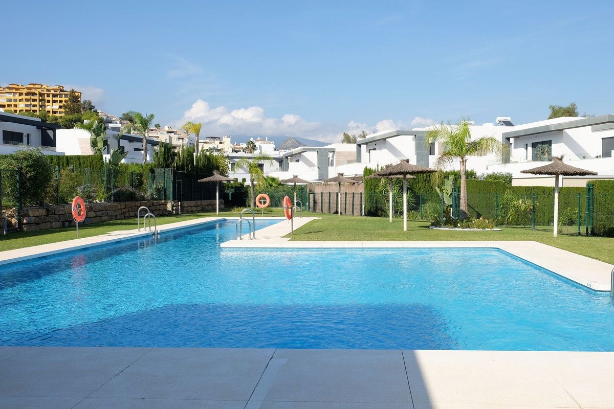 Outdoor swimming pool with sun loungers and umbrellas, surrounded by manicured lawns and modern villas under a blue sky.