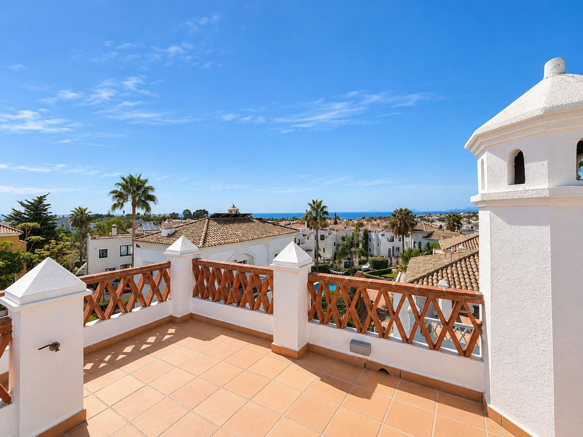 A spacious tiled terrace with white balustrades overlooking rooftops, palm trees, and the distant sea under a clear blue sky.
