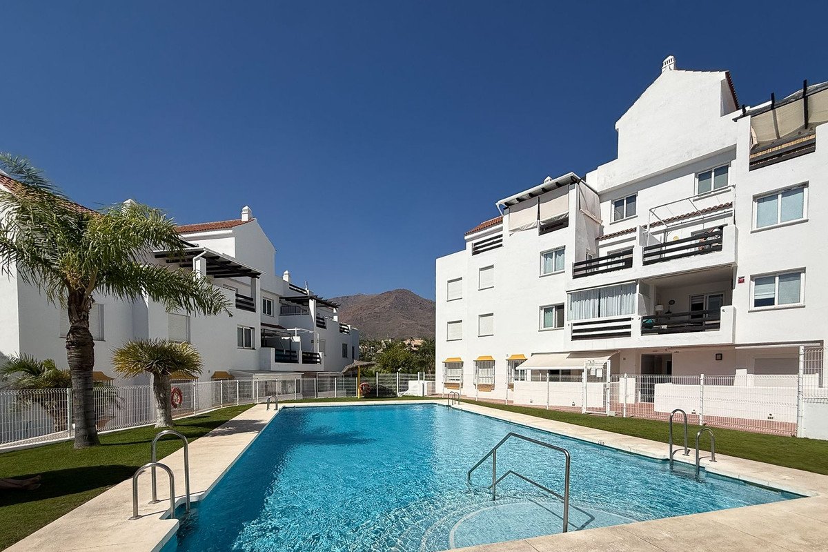 Apartment complex with a swimming pool in the foreground and mountains in the background under a clear blue sky.