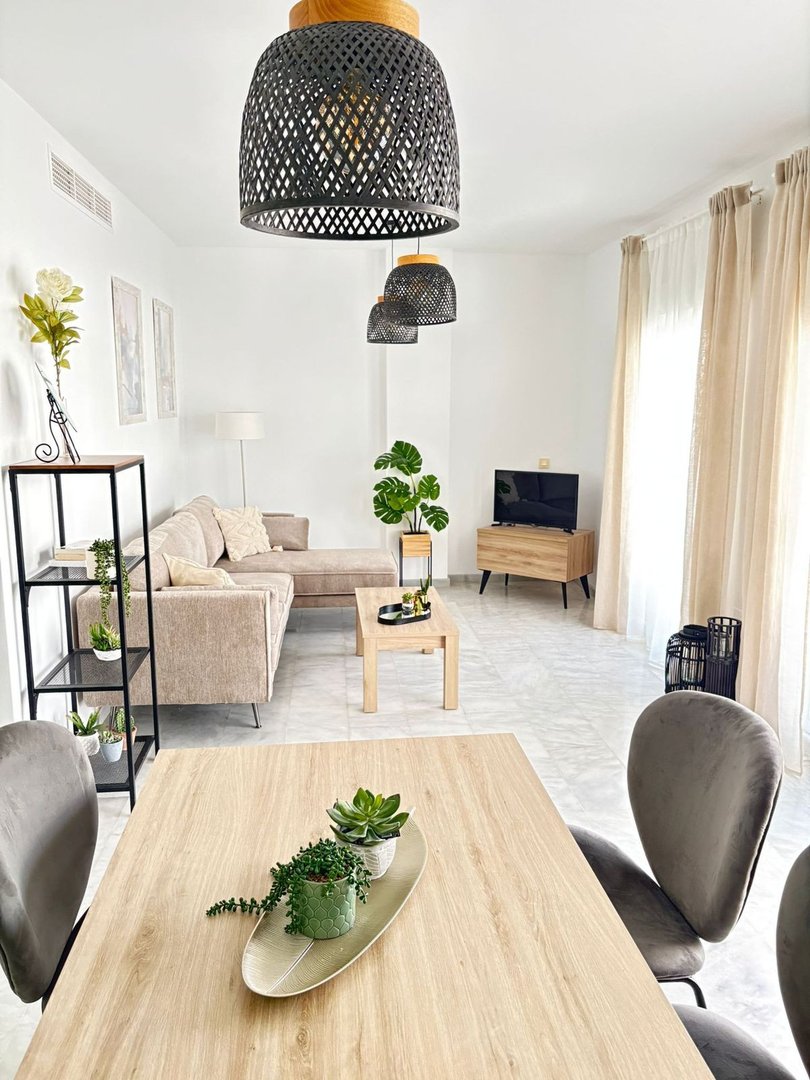 A bright living and dining area with a sectional sofa, coffee table, TV, and dining table with chairs. Natural light streams through the curtains.
