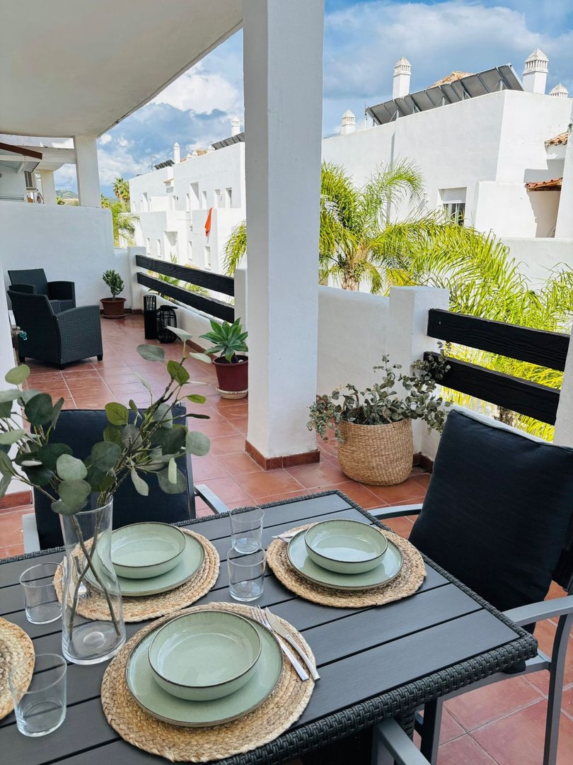 A table set for dining on a terrace with outdoor seating and views of white buildings and palm trees.