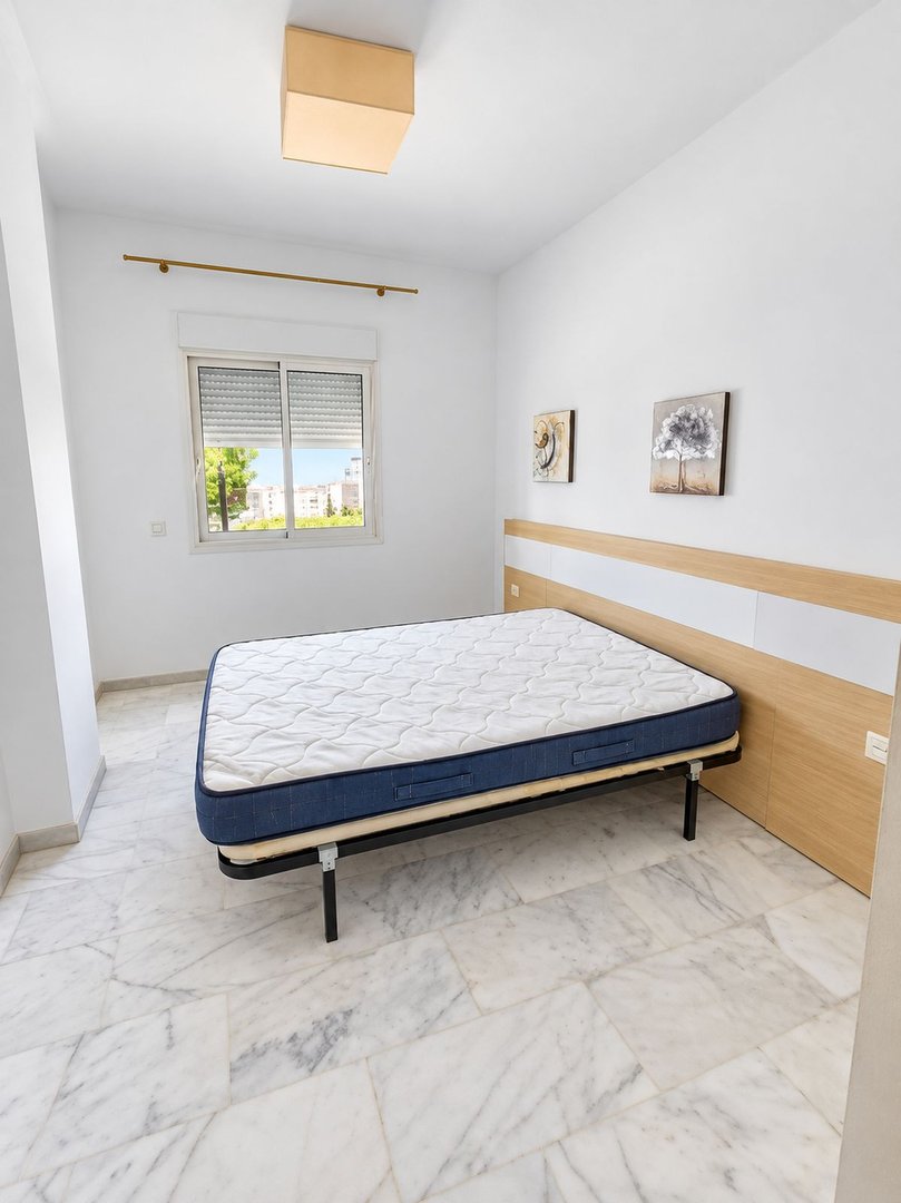 Unfurnished bedroom with a mattress on a frame, a window with a view of buildings and trees, and wall art.