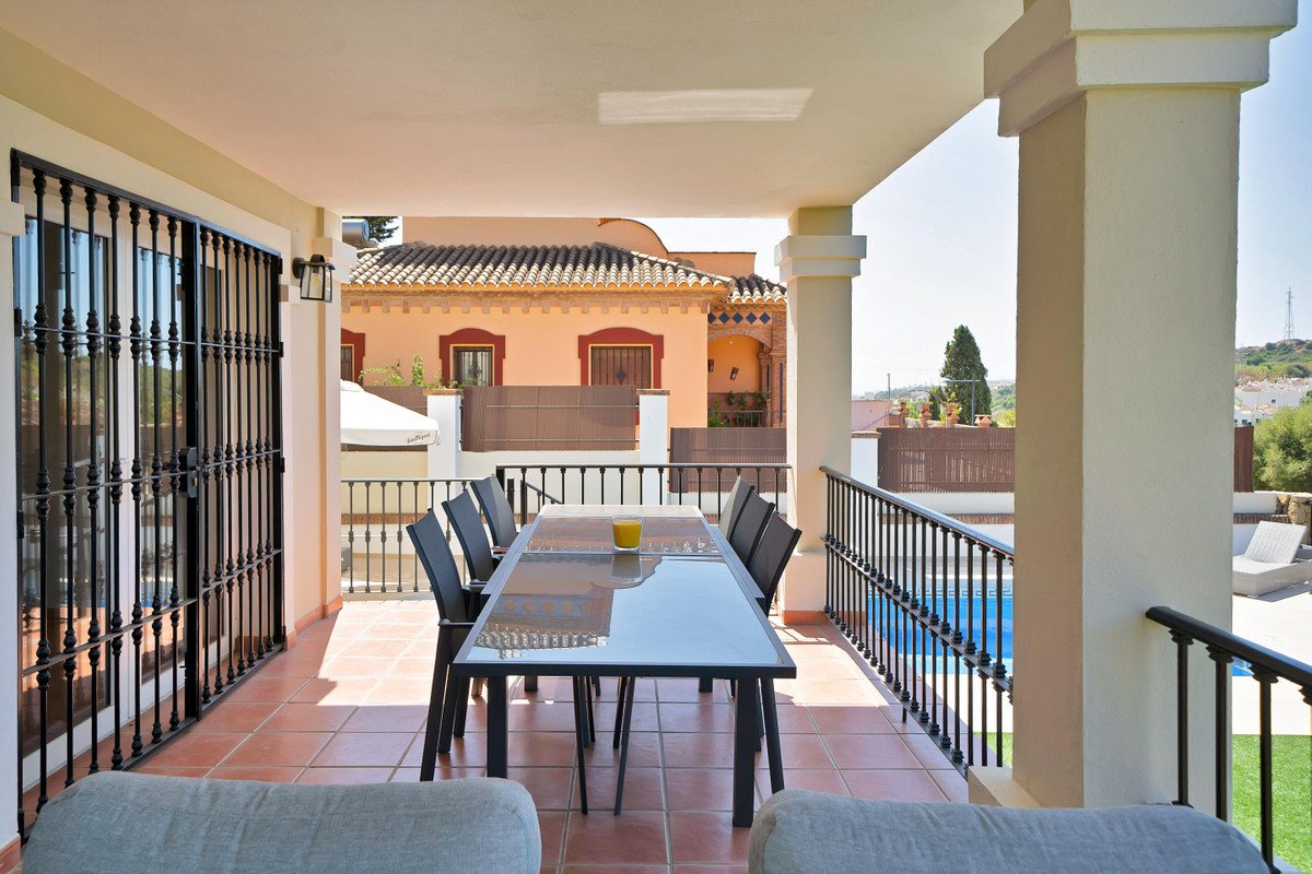 Covered terrace with dining table and chairs overlooking a swimming pool and surrounding buildings.