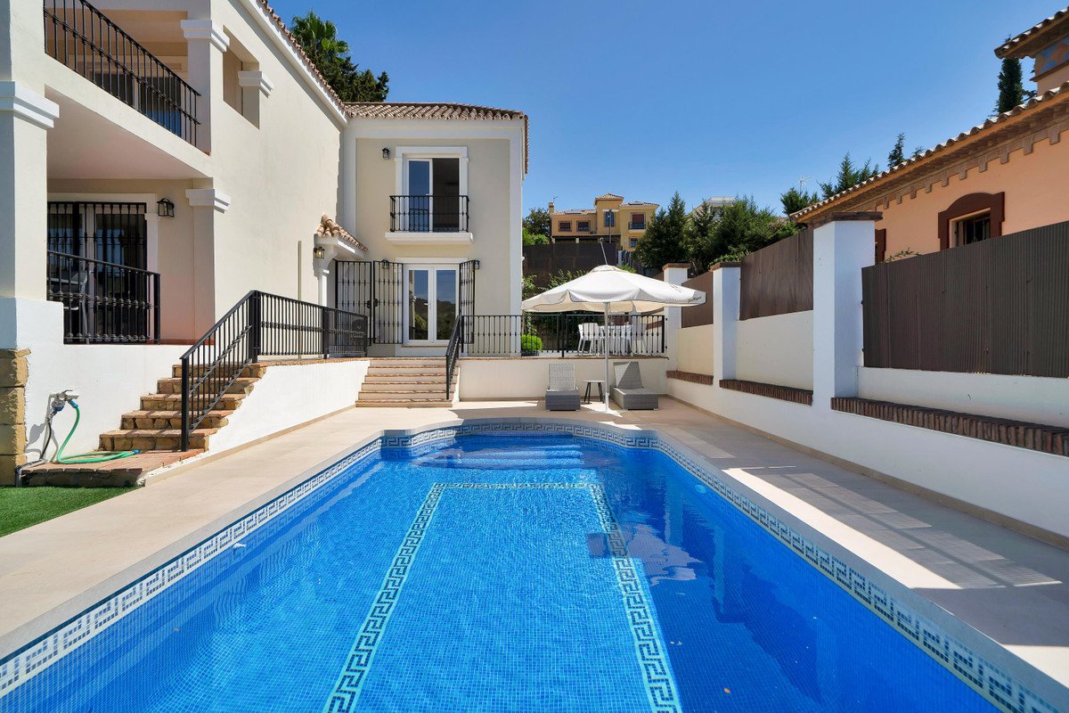 A sun-drenched villa exterior with a bright blue swimming pool, patio, and outdoor seating under an umbrella.