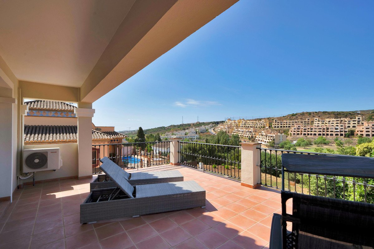 Spacious terrace with lounge chairs, overlooking a pool and a hillside town with distant hills under a clear blue sky.