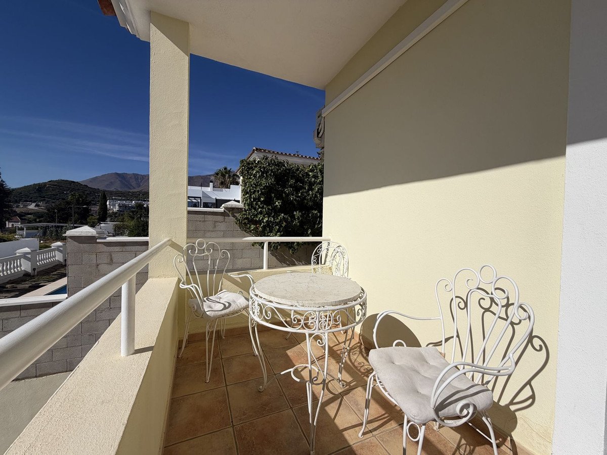A sunny balcony with white metal table and chairs, overlooking a landscape with mountains and houses under a clear blue sky.