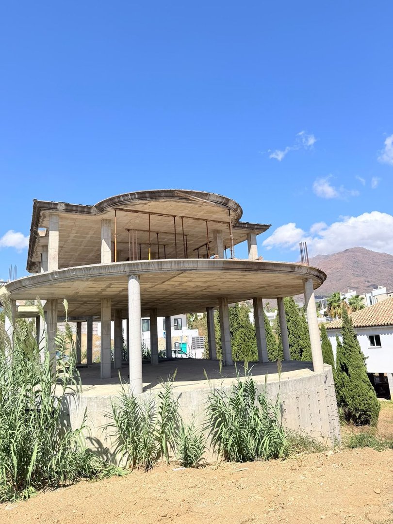 Under construction modern house with multiple levels and columns, surrounded by greenery and mountains in the background.