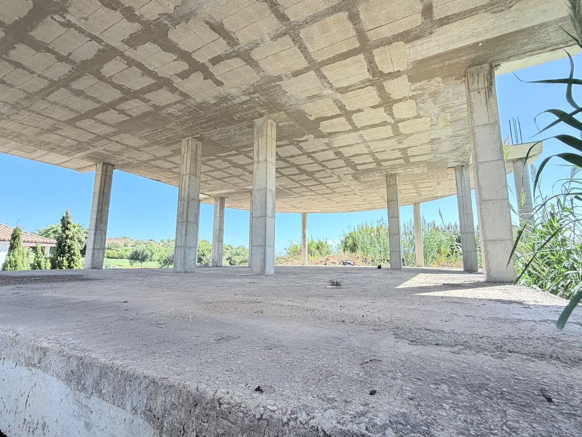 Under-construction concrete balcony with pillars and surrounding greenery, offering a scenic countryside view under a clear blue sky.