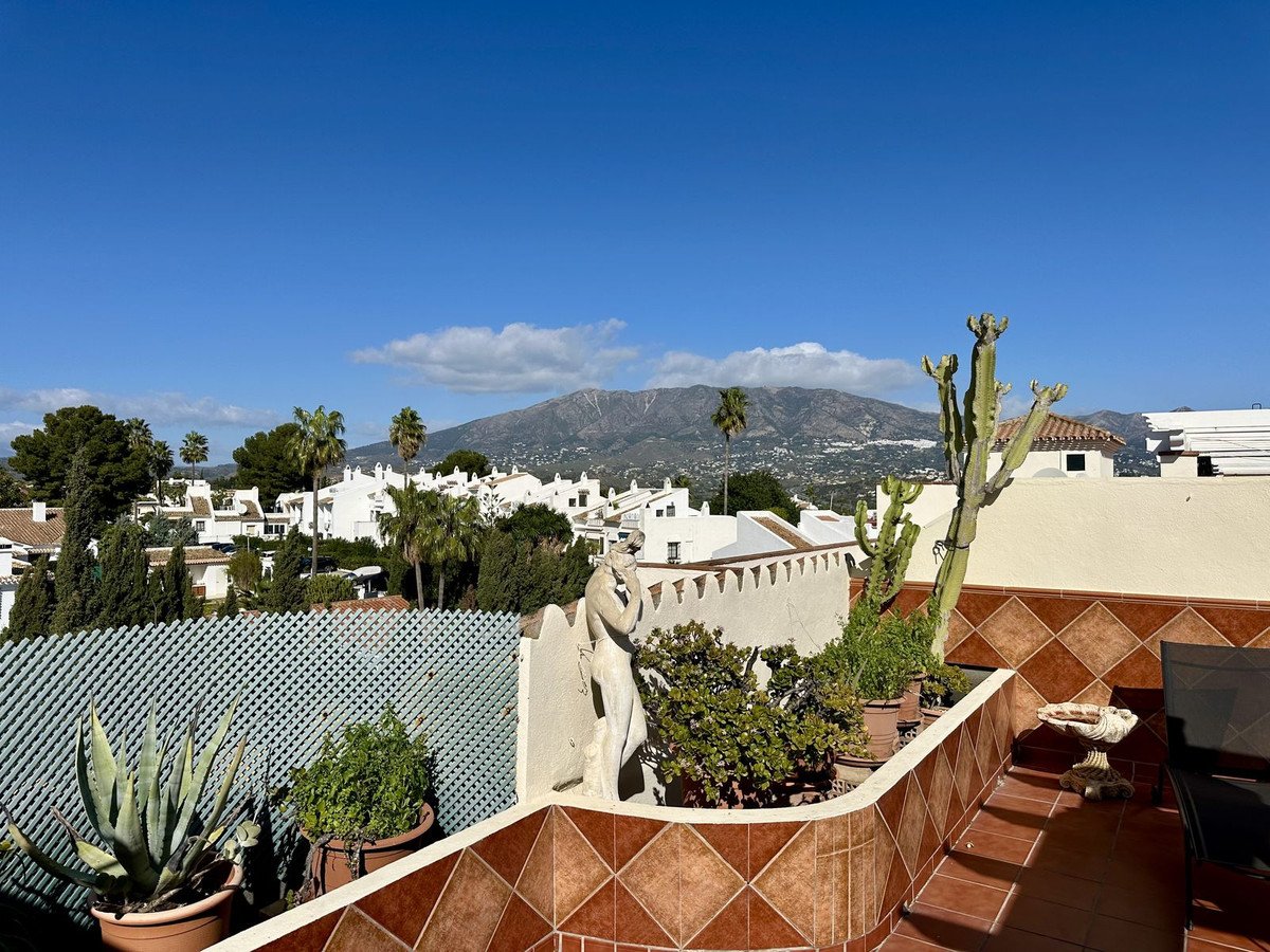 Terrace with potted plants and a statue, overlooking white houses and mountains under a clear blue sky.