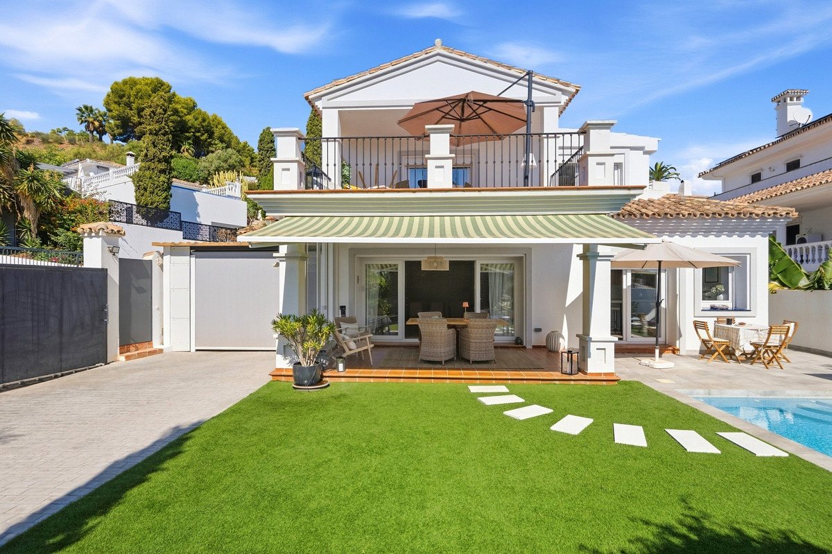 Villa with a covered terrace, outdoor dining area, green lawn, swimming pool, and balcony with umbrella.