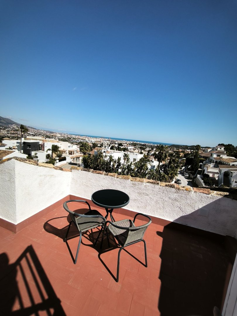 Terrace with two chairs and a table, overlooking a town and the sea under a clear blue sky.